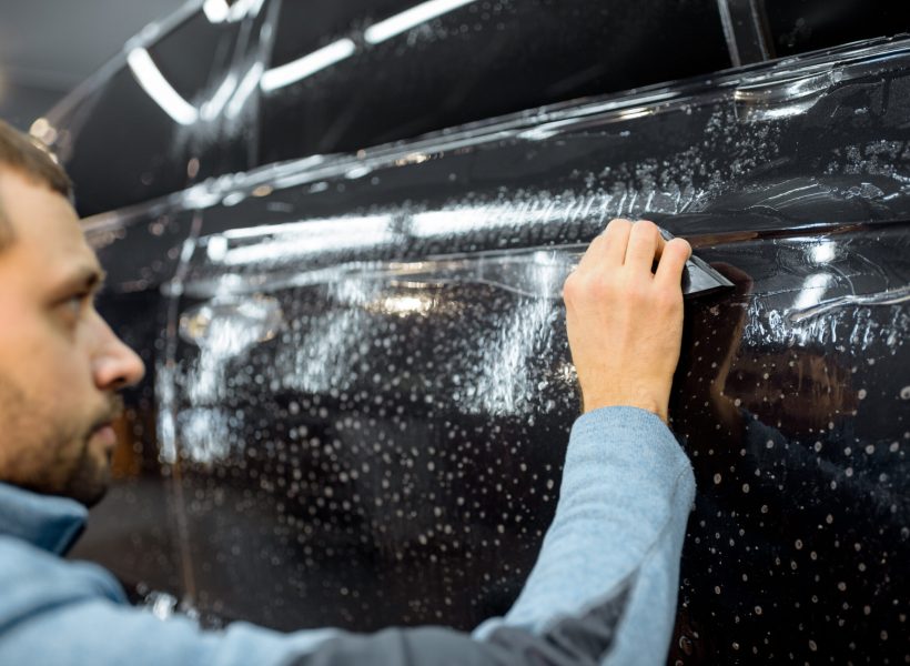 Car service worker sticking anti-gravel film on a car body with scrapper at the detailing vehicle workshop. Concept of car body protection with special films