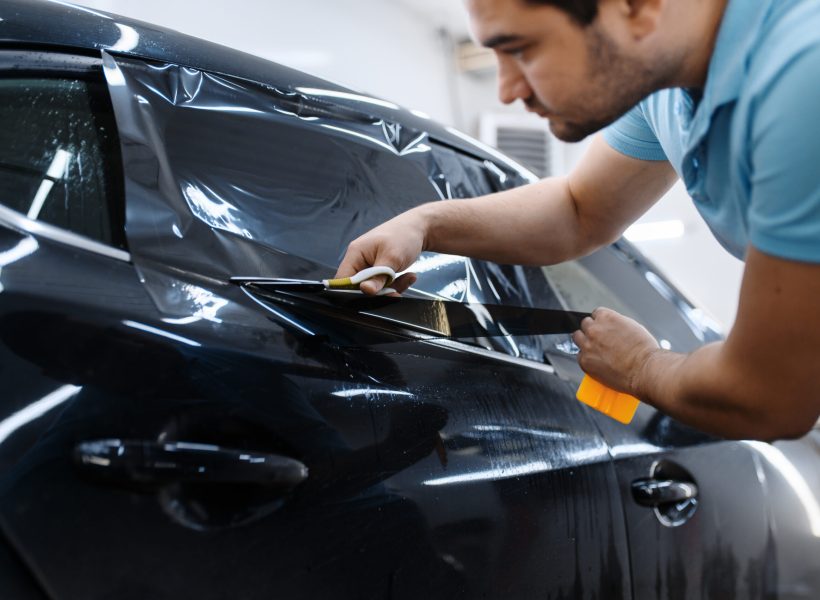 Male worker holds sheet of film, car tinting installation, tuning service. Mechanic applying vinyl tint on vehicle window in garage, tinted automobile glass
