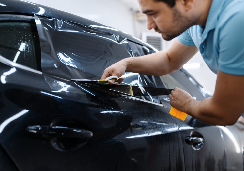 Male worker holds sheet of film, car tinting installation, tuning service. Mechanic applying vinyl tint on vehicle window in garage, tinted automobile glass
